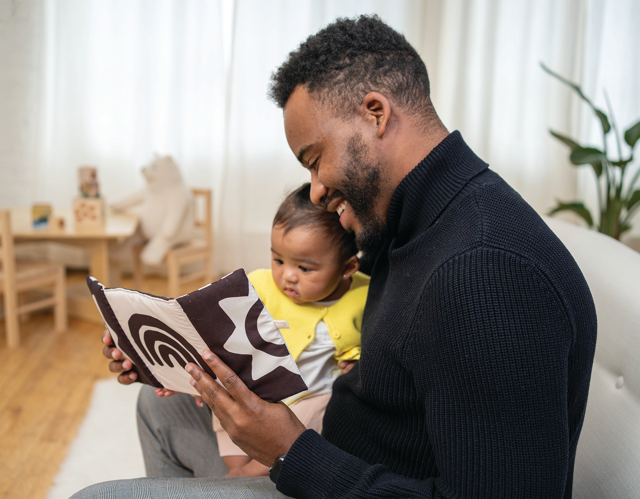 Dad and baby seated on couch playing with a soft peek-a-boo mirror toy